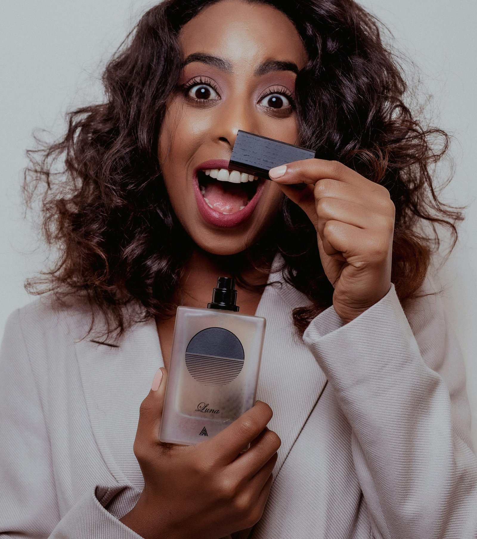 Joyful woman with curly hair posing with luxury perfume in a studio setting, showcasing excitement and beauty.